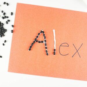 Children spell their name written on a piece of paper using liquid glue and small black beans.